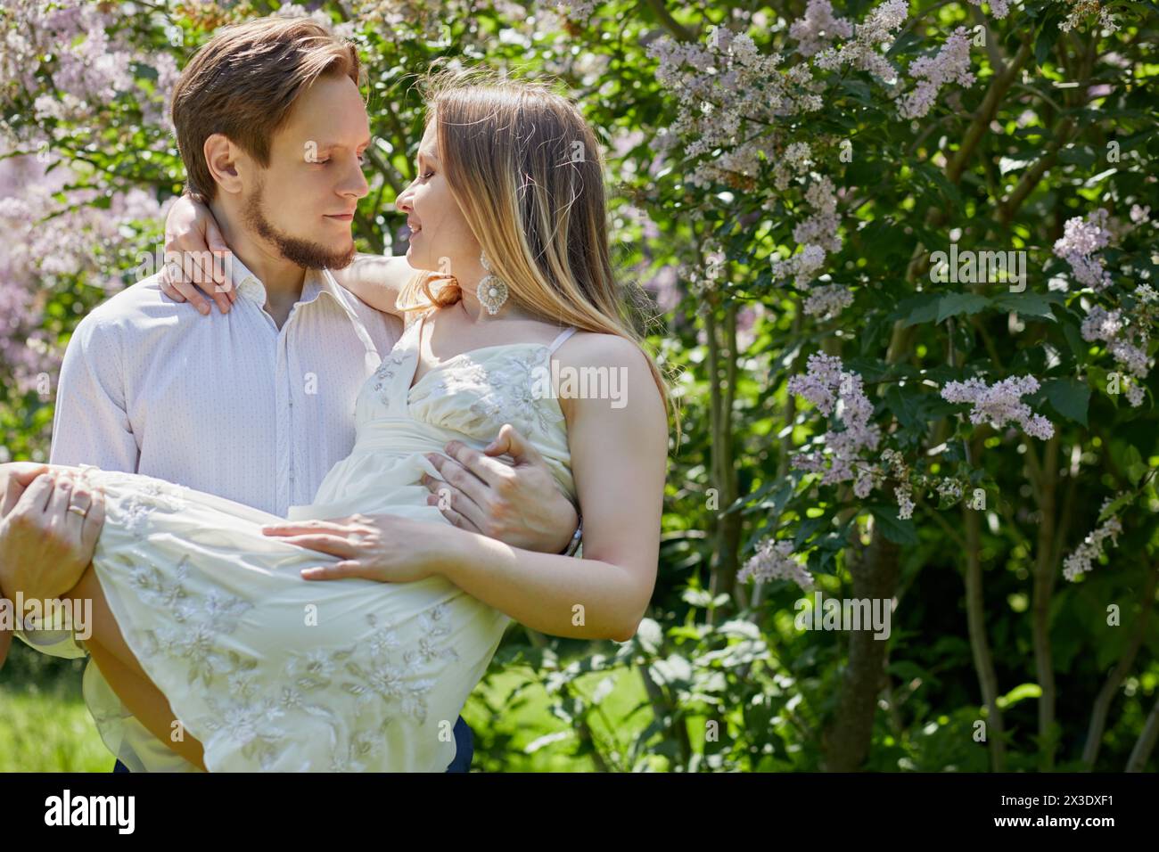 Young bearded man holds on arms woman in park with blooming bushes and trees. Stock Photo