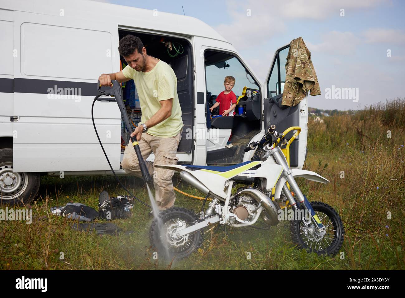 Man washes motorbike after training outdoor near minibus, little son looks at him from driver cabin. Stock Photo