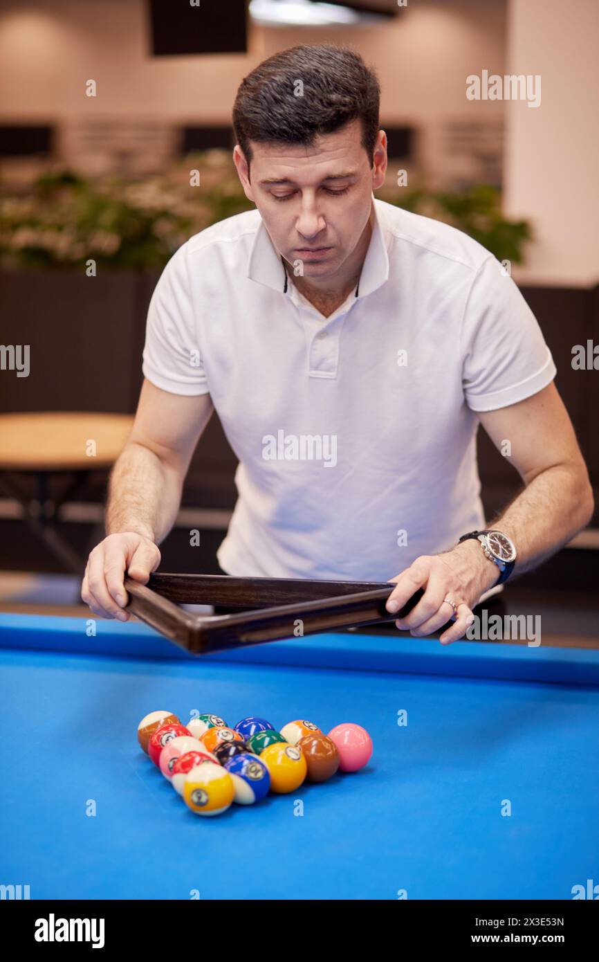 Man in white polo-neck shirt arranges pool balls on table in club. Stock Photo