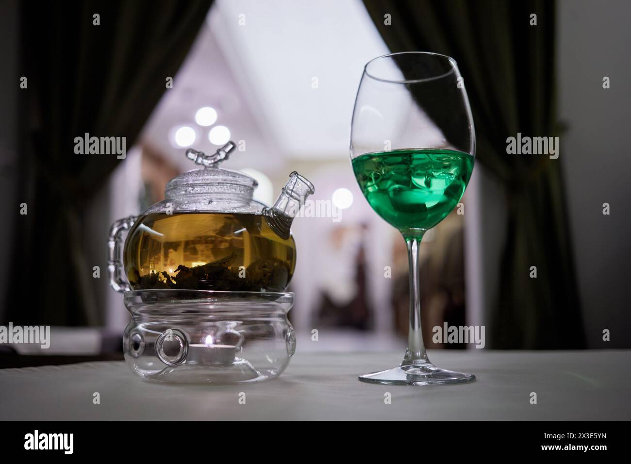 Transparent glass teapot on candle heater and wineglass with green beverage with ice on table. Stock Photo