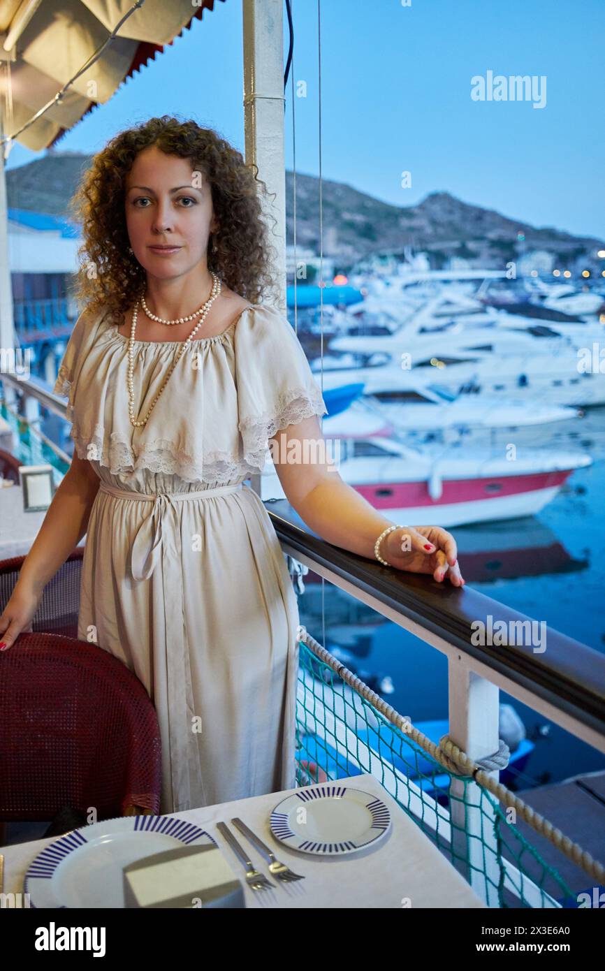 Smiling curly woman stands leaning on chair back and railing in floating cafe with bay view. Stock Photo