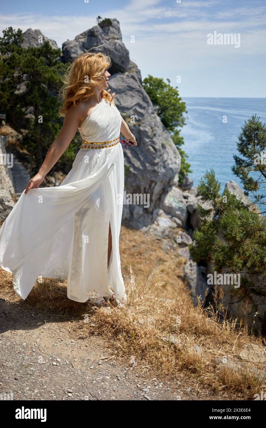 Smiling blond woman in white dress with golden waist belt stands on cliff and looks at sea on sunny day. Stock Photo