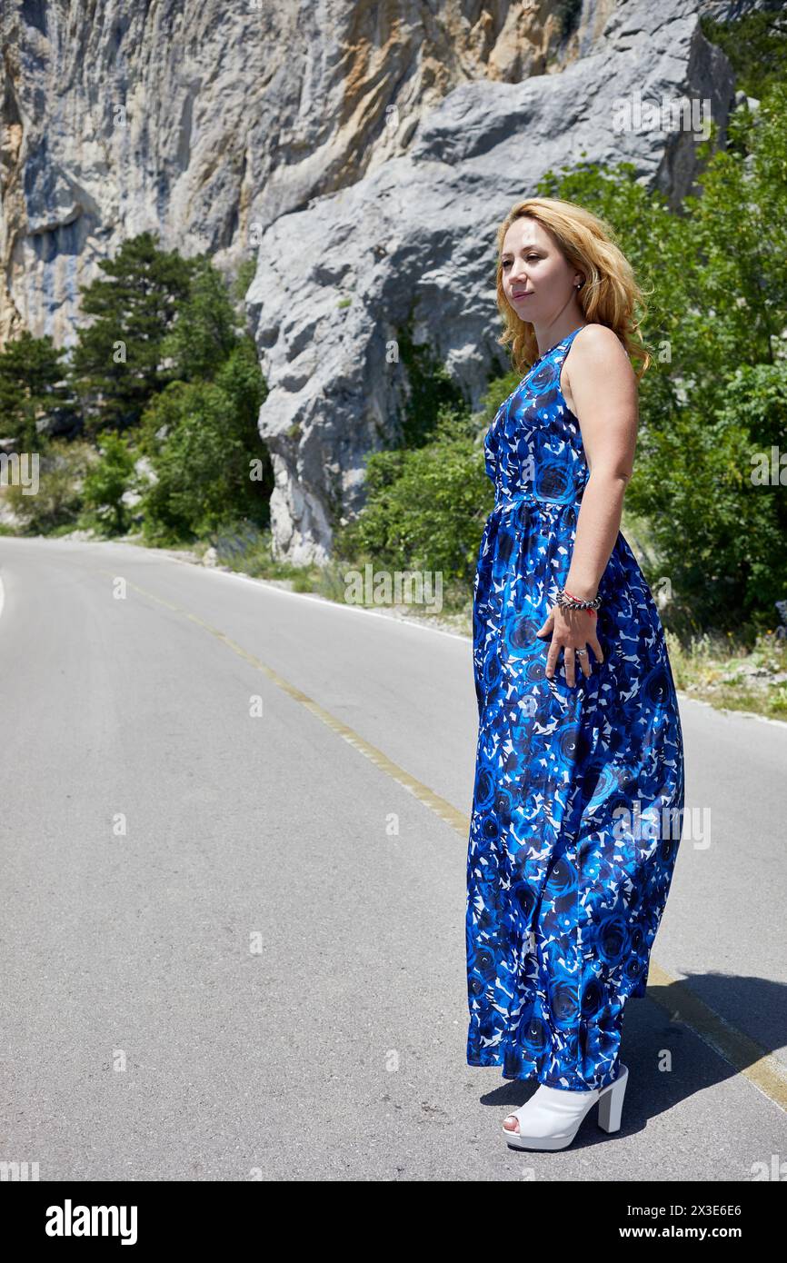Blond woman in blue dress and white high-heel shoes stands on road under cliff in mountains. Stock Photo
