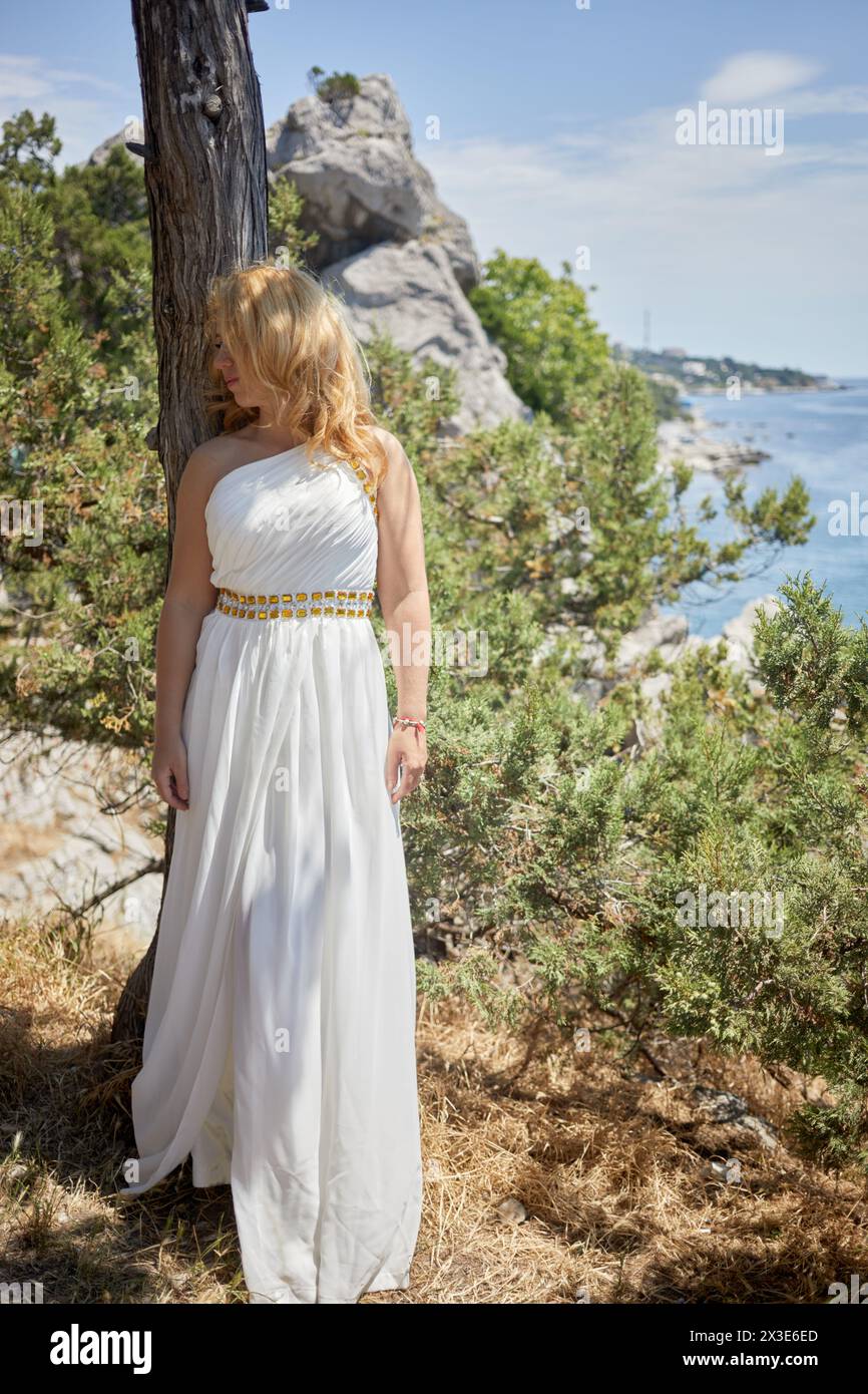 Blond woman in white dress with golden waist belt stands leaning her back to tree trunk with cliff and seashore at background on sunny day. Stock Photo