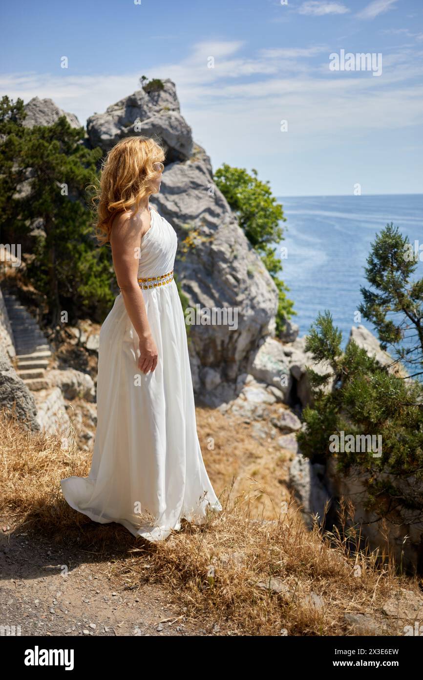 Blond woman in white dress with golden waist belt stands on cliff looking at sea on sunny day. Stock Photo