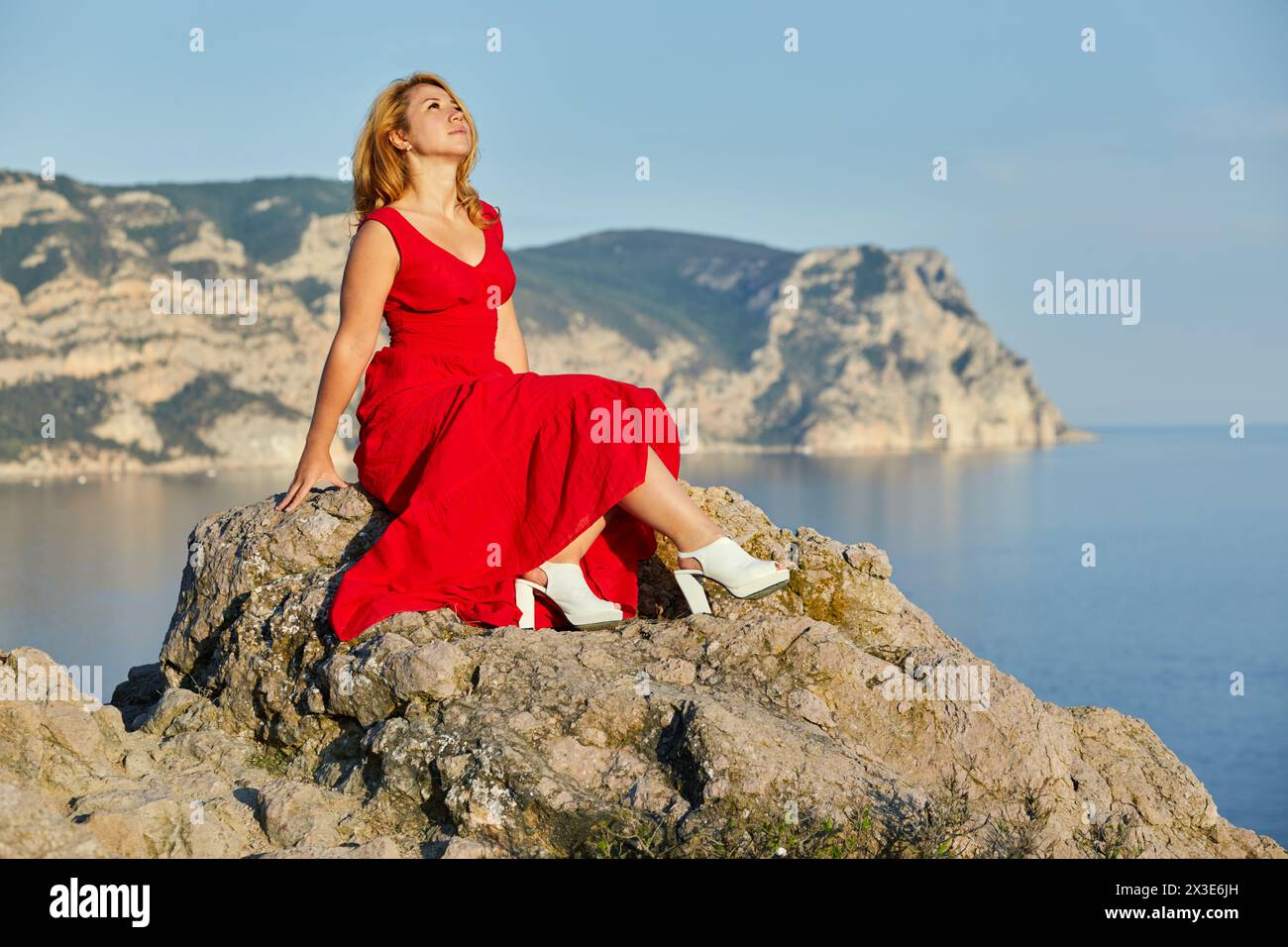 Blond woman in red dress and white high-heel shoes sits on top of rock against hills, sky and sea. Stock Photo