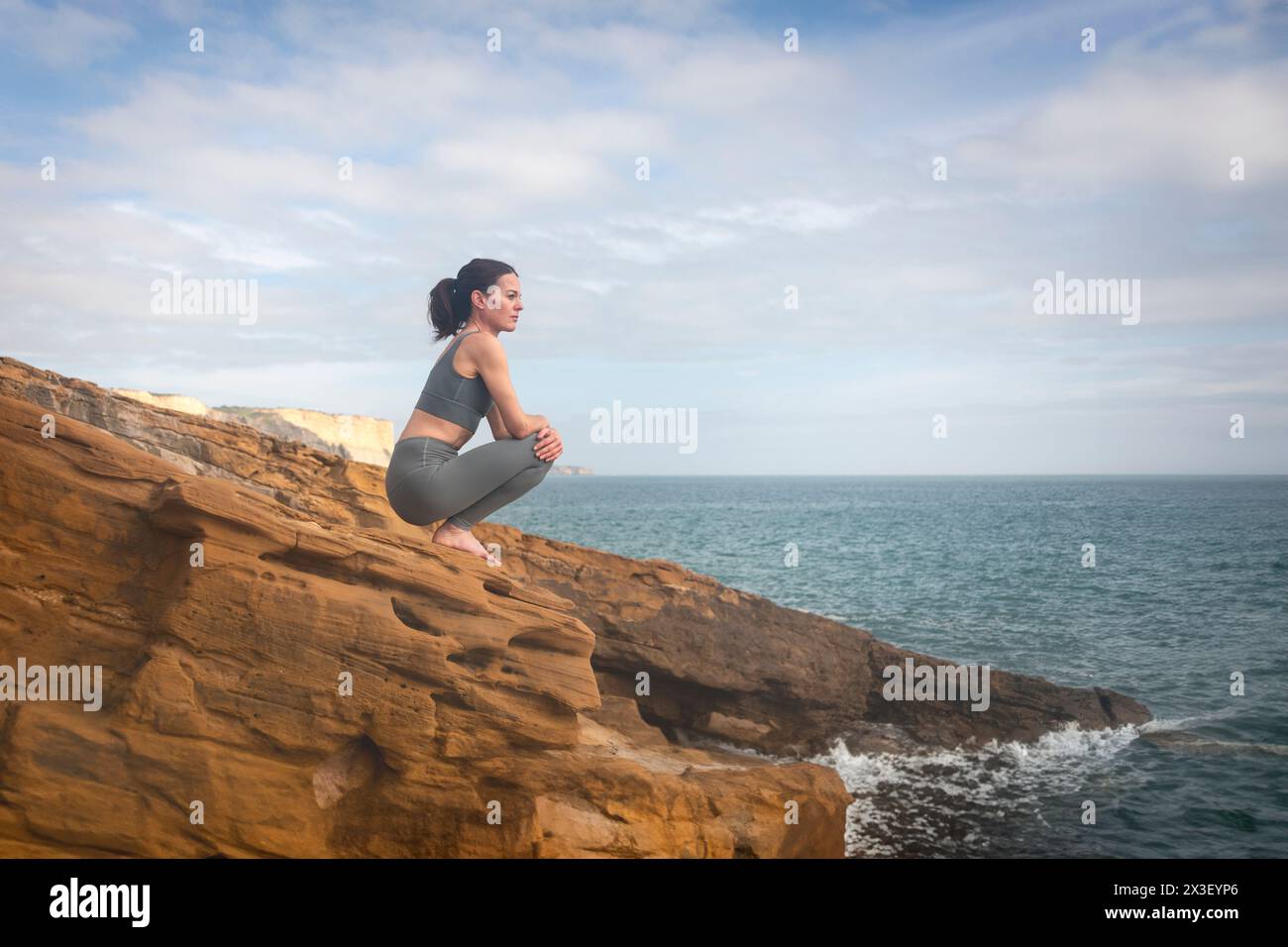 Sporty woman sitting on a rock looking out to sea, de-stressing after workout or yoga Stock Photo
