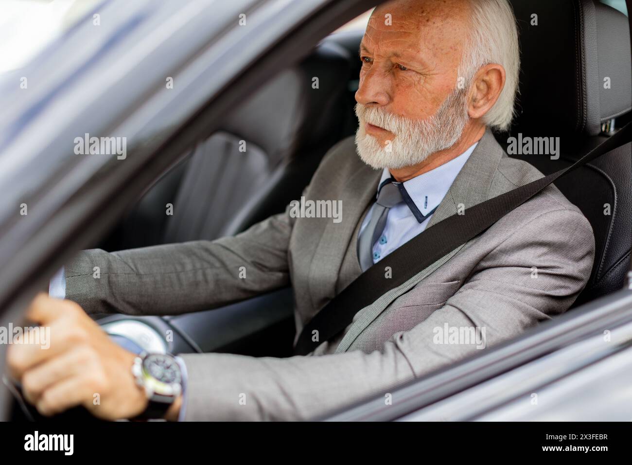 Cheerful elderly man in a suit driving leisurely, basking in the warm glow of a bright day Stock Photo