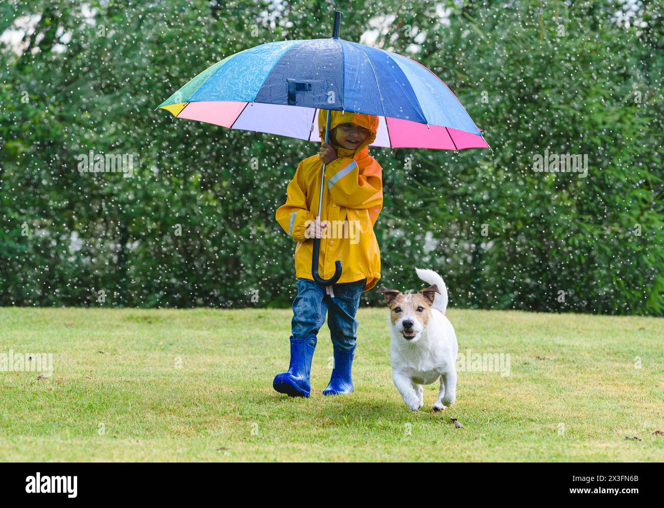 Kid in raincoat under umbrella and her dog playing on grass lawn during spring rain Stock Photo