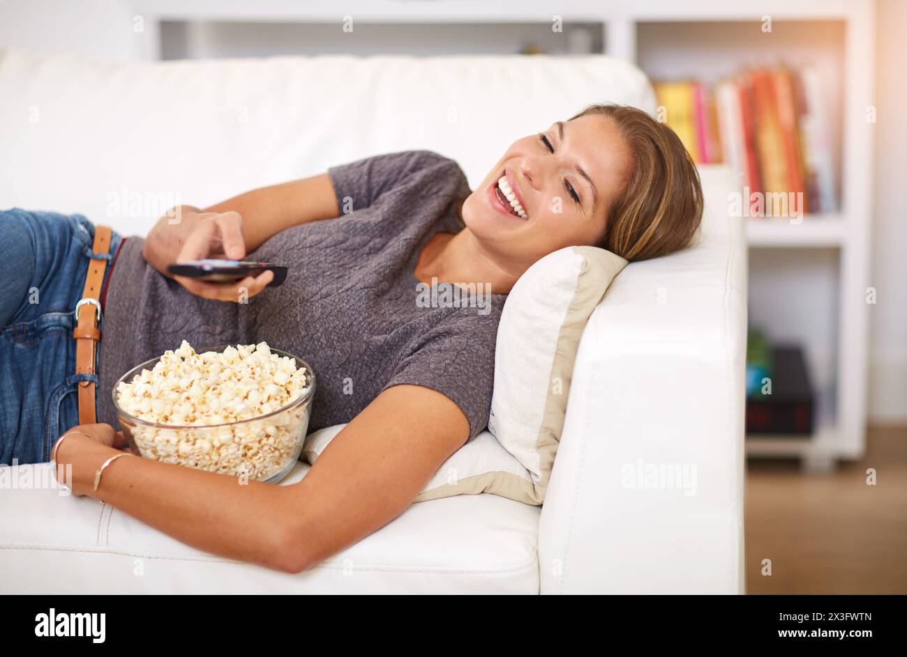 Woman, sofa and popcorn with remote and laugh in home, snack and watching on weekend. Comedy, entertainment and food for movie or show, streaming or Stock Photo