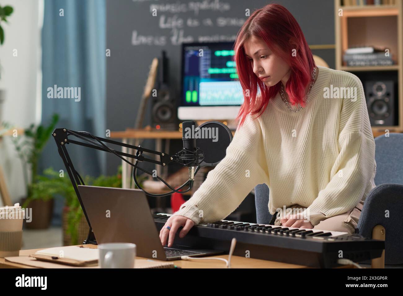 Cute teenage girl bending over desk with electric piano and laptop and looking at device screen while pressing key during sound recording Stock Photo