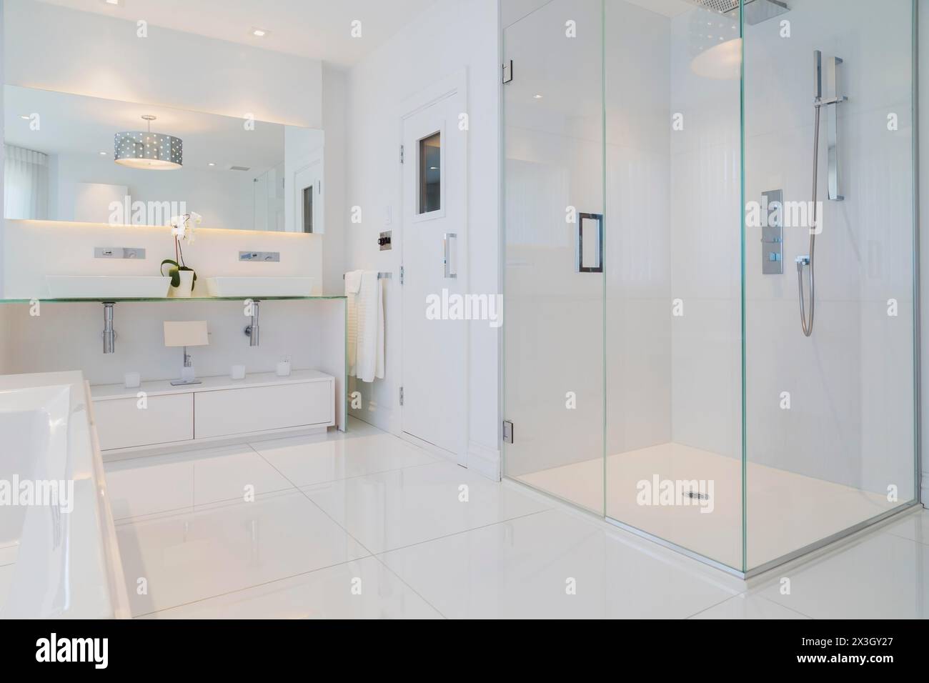 Partial view of white elongated rectangular bathtub and cabinets with large wall mirror, glass shower stall, sauna room door in en suite with ceramic Stock Photo