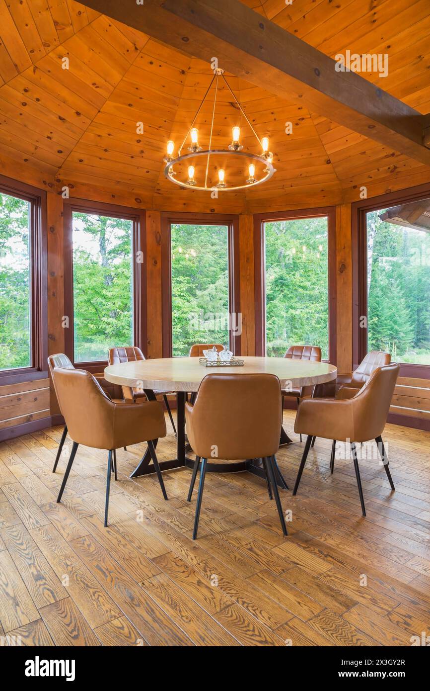 Round wooden dining table with tan leather armchairs in dining room inside a milled Eastern white pine timber and flat log profile home, Quebec Stock Photo
