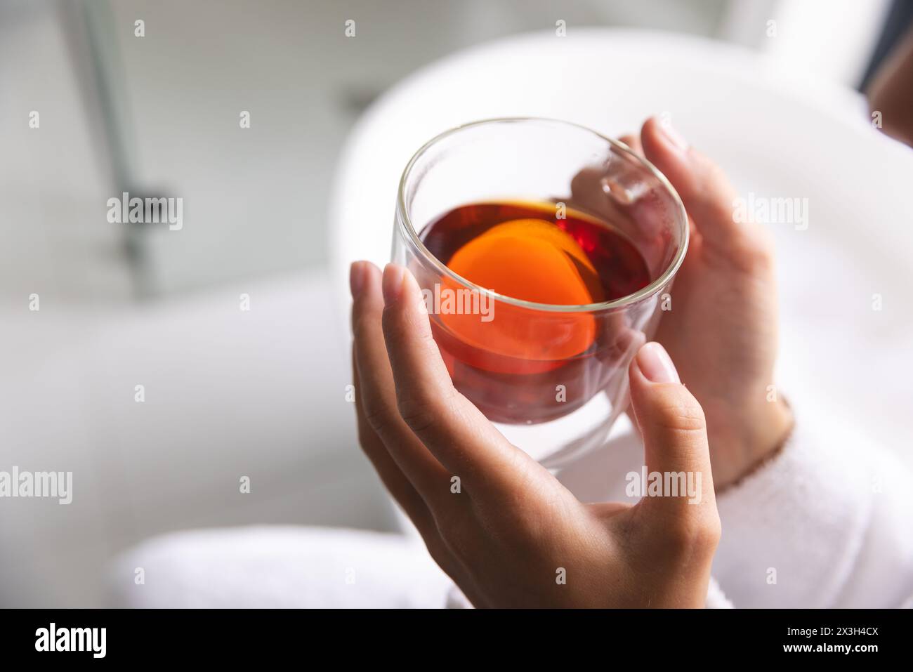 At home, young Caucasian woman holding a glass of tea wears a white robe Stock Photo