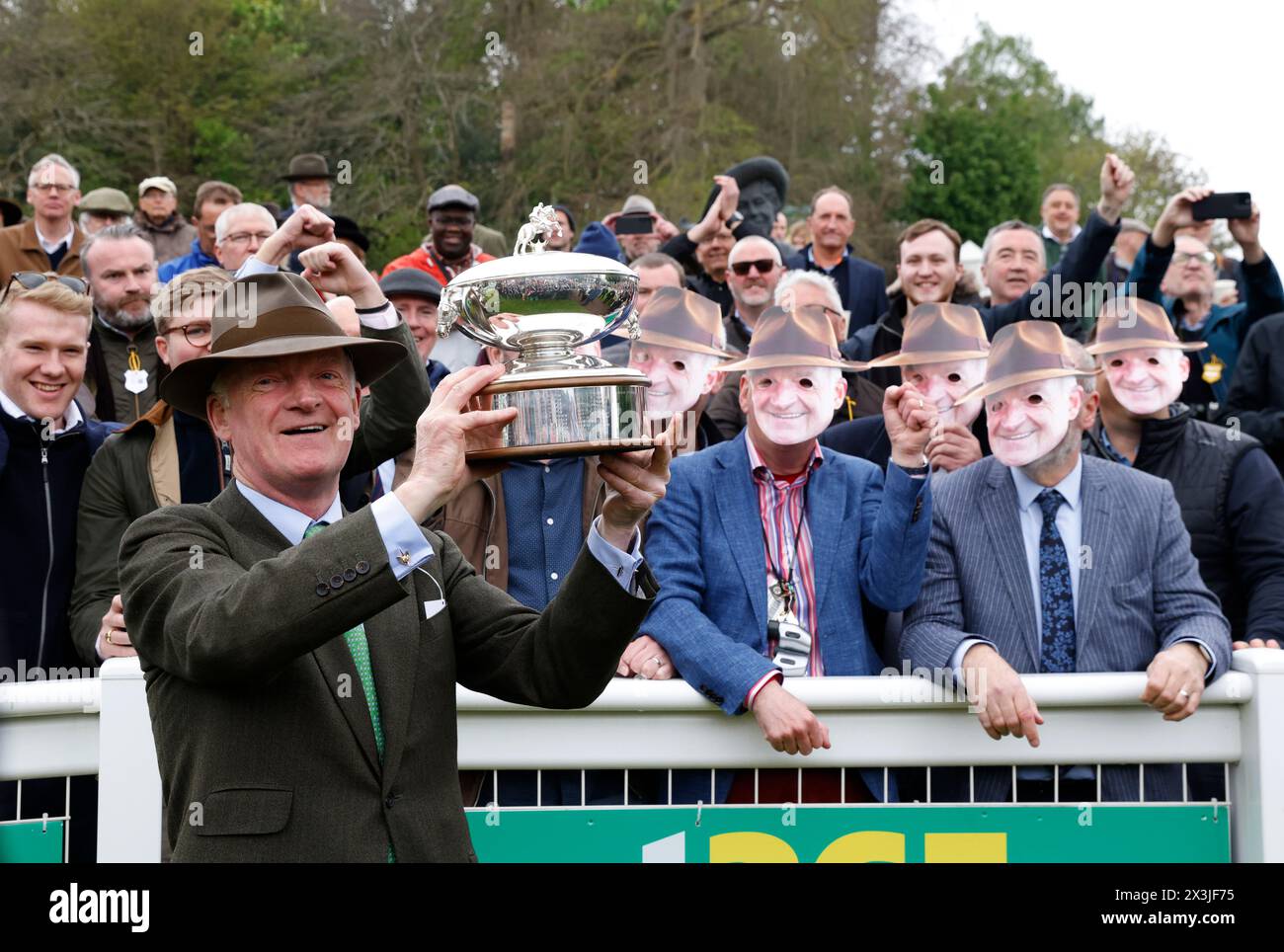 Winning trainer Willie Mullins with the Champion Trainer trophy poses with racegoers wearing masks of his face after Minella Cocooner won the bet365 Gold Cup Handicap Chase on bet365 Jump Finale day at Sandown Park Racecourse, Esher. Picture date: Saturday April 27, 2024. Stock Photo