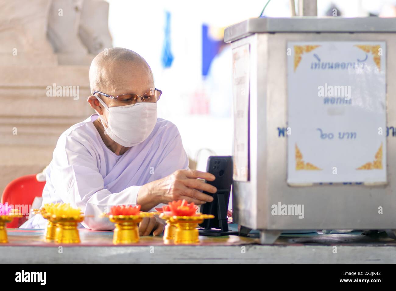 SAMUT SAKHON, THAILAND, MAR 24 2024, Buddhist nun is looking at mobile phone on table with box for donated money, inside Buddhist temple Stock Photo
