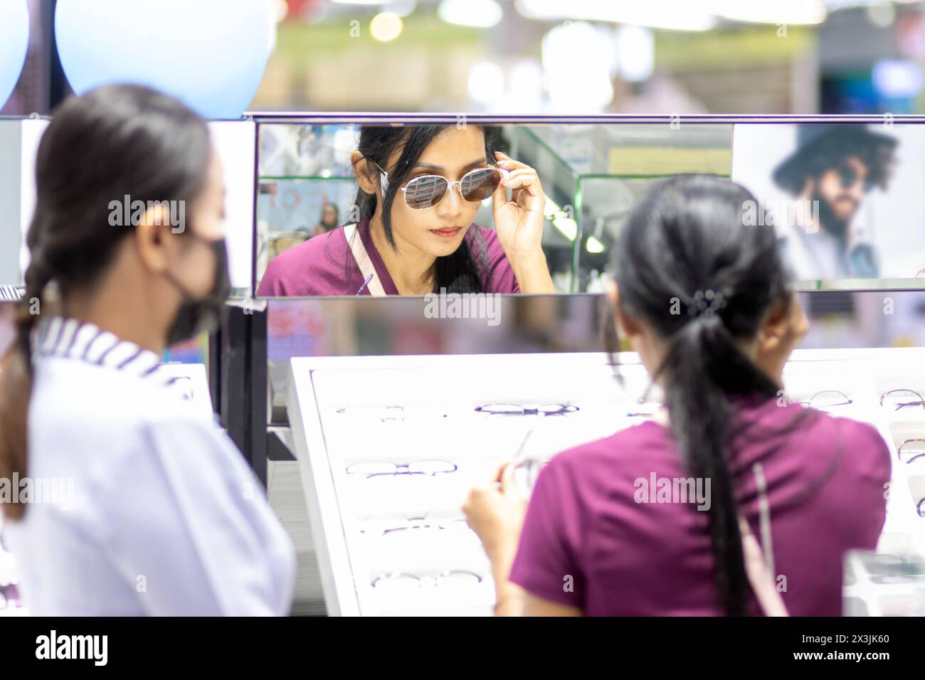 A young woman tries sunglasses at the mirror in an optical shop with the assistance of a saleswoman Stock Photo