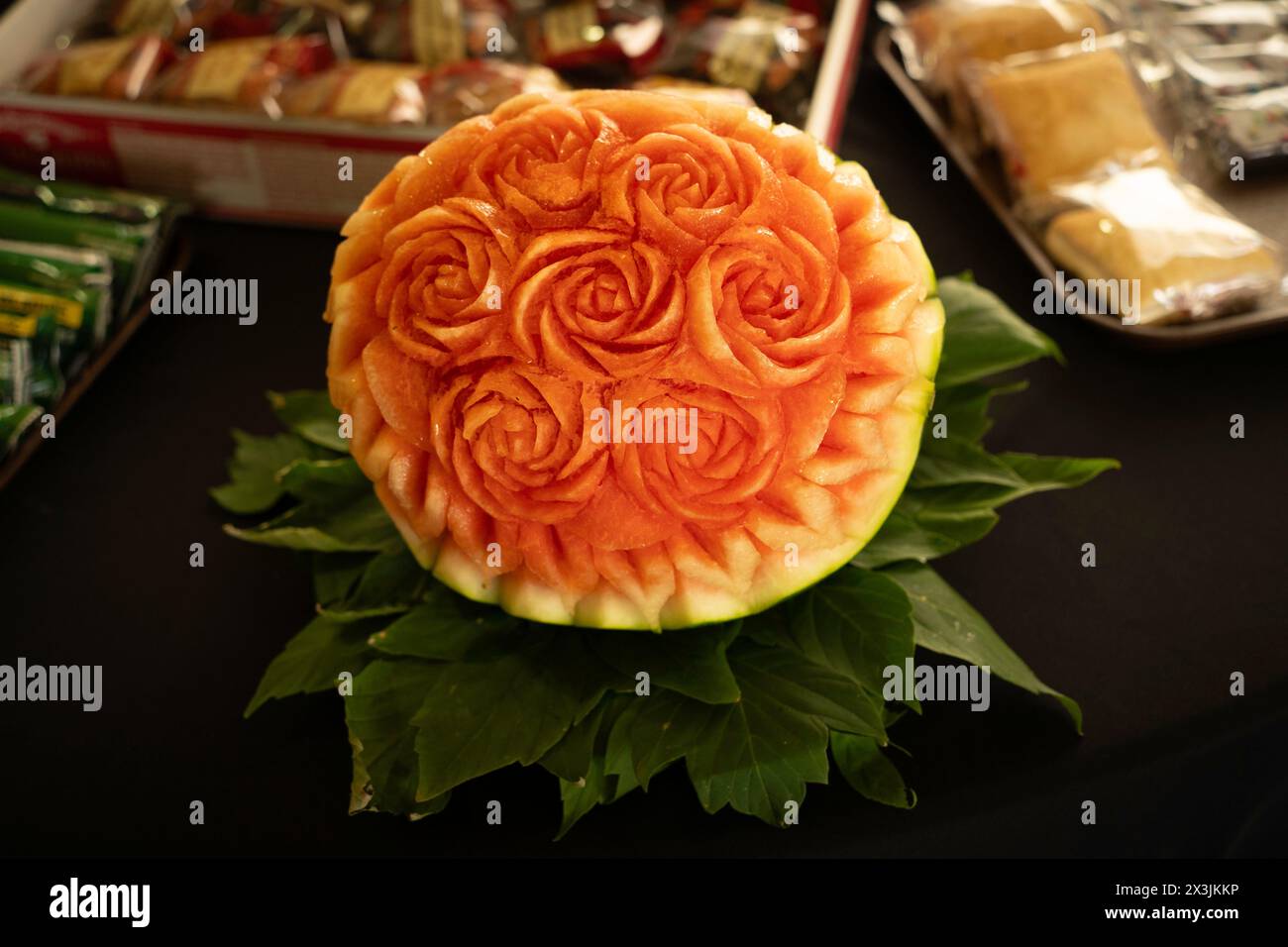 A watermelon carving of a boquet of roses sits on a black table with package snacks placed around it. Stock Photo