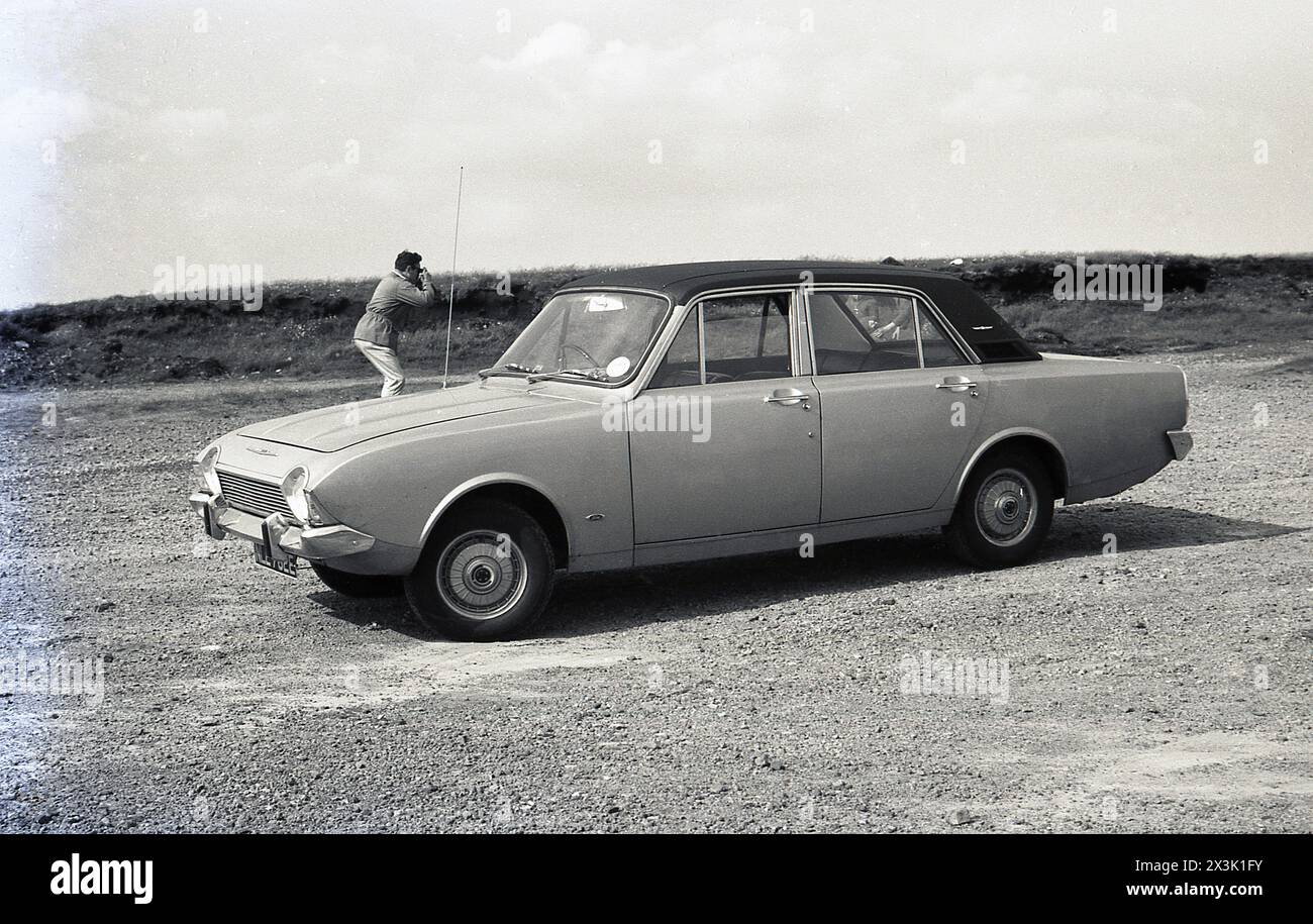 1970s, historical, a Ford Corsair motorcar parked outside on rough ground, the male owner outside taking photograph. The car was produced from 1964 - originally known as the Ford Consul Corsair, later simply the Ford Corsair - until 1970, with different variations. The model here is a Corsair V4 2000E, a vinyl roofed Corsair, designed to compete with other more upmarket executive cars such as the Rover 2000 and Triumph 2000. Stock Photo