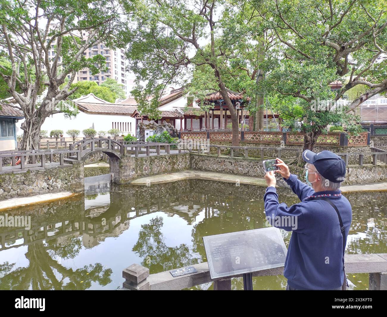 Banqiao District, Taipei, Taiwan, March 27, 2024: Elderly asian man with mask takes photo in Lin Garden in Taipei of pond and bridge with smartphone Stock Photo