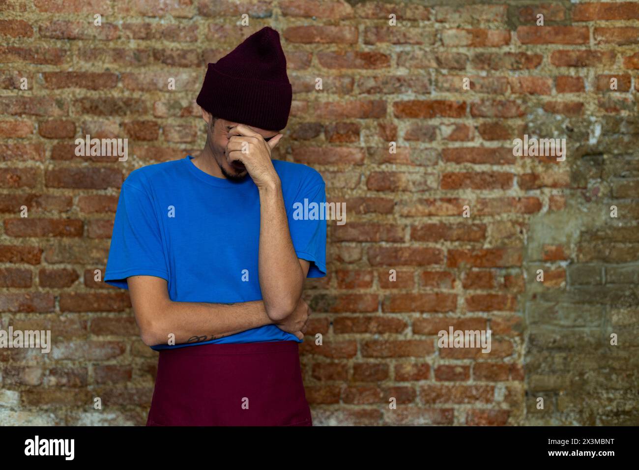 l Young Latino man (28) covers his face out of embarrassment. Copy space, brick background. Stock Photo