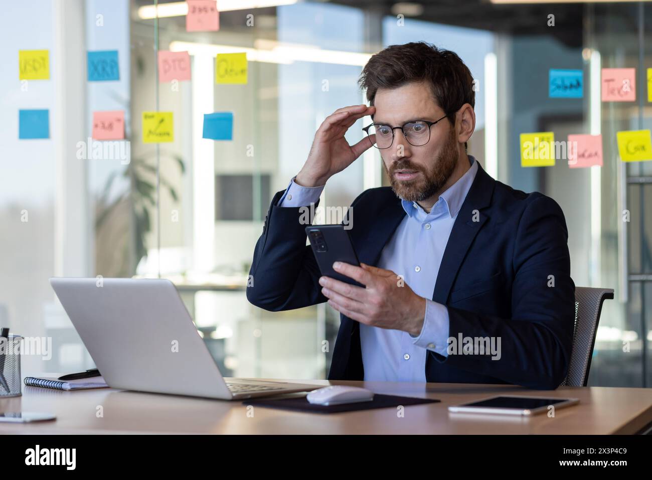 A man in a suit is looking at his cell phone while sitting at a desk. The scene suggests that he might be working or attending to some important matter. The man's facial expression Stock Photo