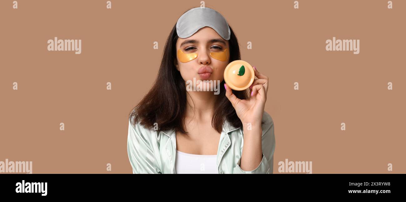 Grimacing young woman with under-eye patches holding jar of cosmetic cream on brown background Stock Photo