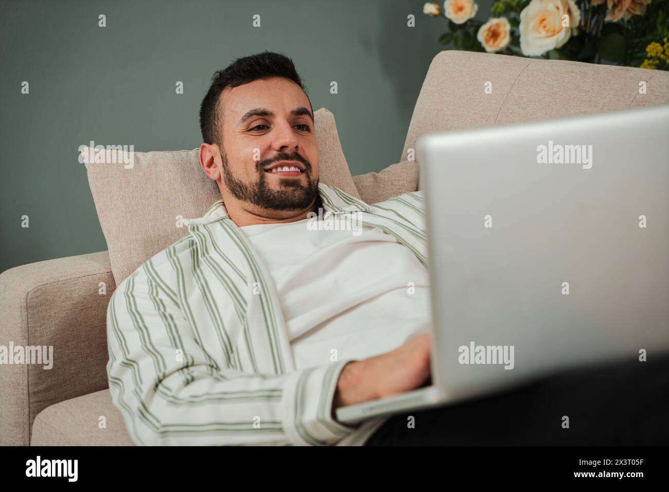 Handsome Young Adult Man Using Laptop While Sitting On Couch At Home. Smiley Millennial Guy Working Remotely On Computer Laying On A Sofa At Cozy Apartment Living Room. Happy Male Enjoying Online Job. High quality photo Stock Photo