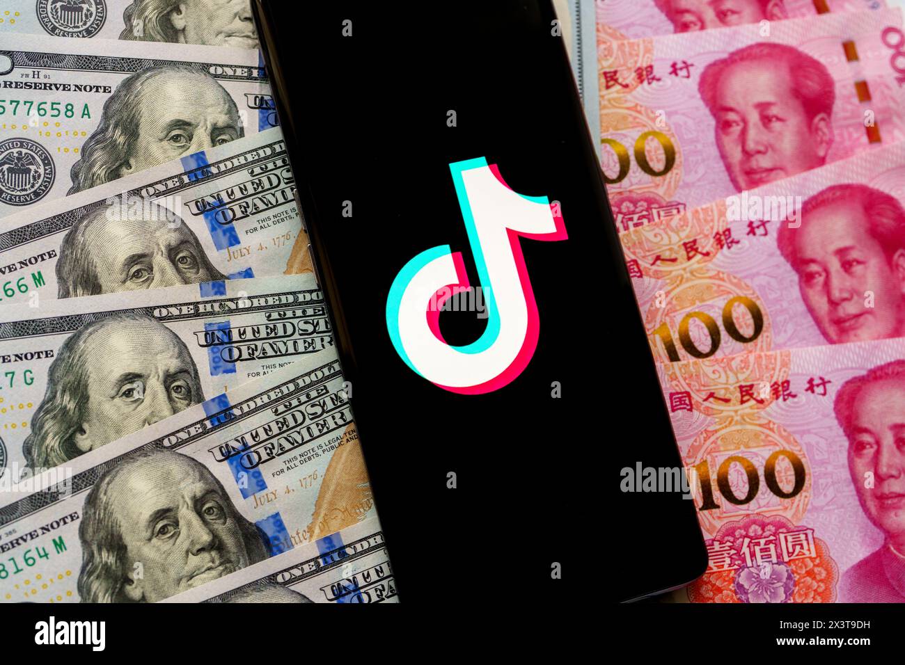 TikTok app logo seen on the screen of smartphone, which is placed on US dollars an Chinese Yuan banknotes. Stafford, United Kingdom, April 28, 2024 Stock Photo