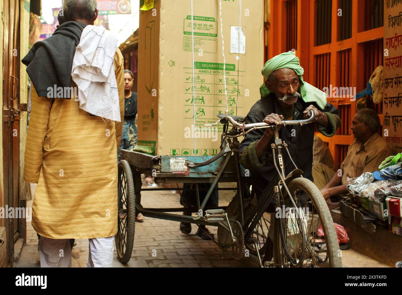 Elderly man pushing a cycle goods carrier loaded with a refrigerator through an alleyway in Varanasi, India. Stock Photo
