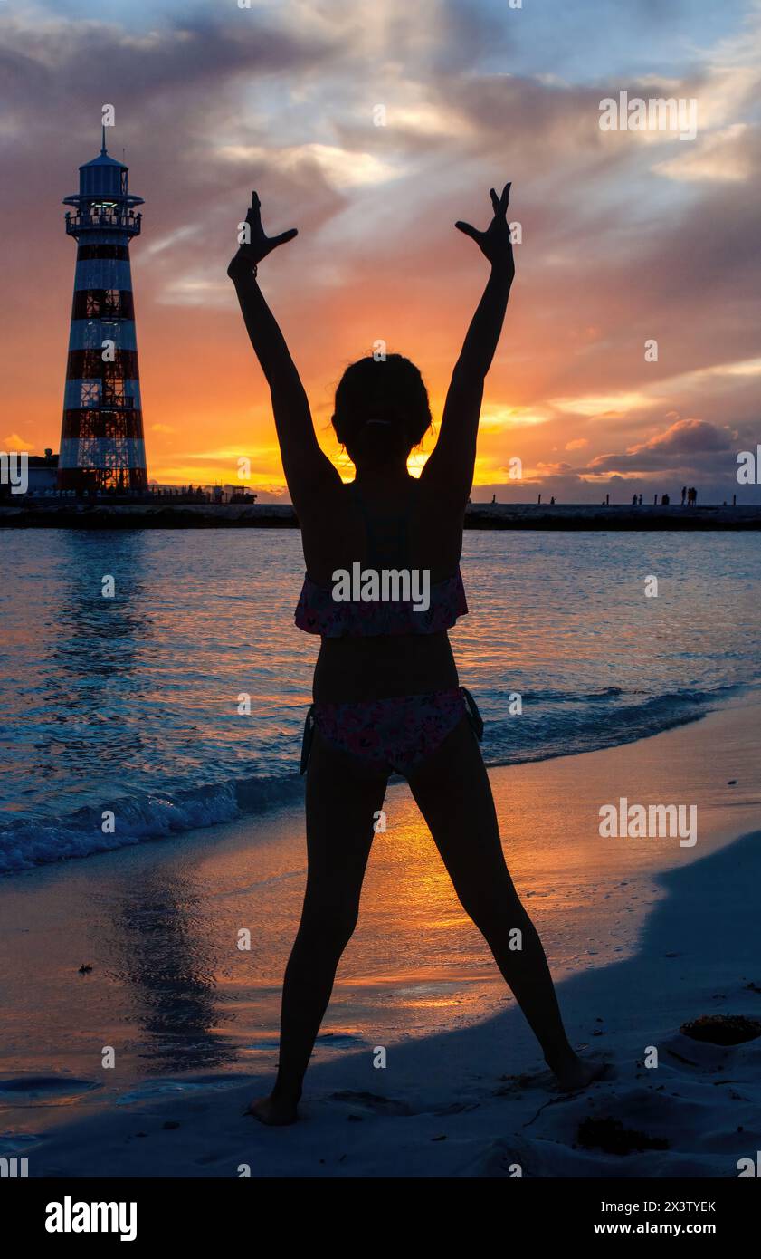 Backlit silhouette of a young woman looking towards the lighthouse at sunset on a Bahamian beach. Stock Photo