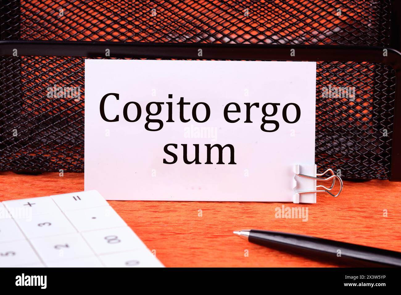 The words Cogito Ergo Sum or I think Therefore I Am on a clean white business card next to a calculator, a pen on an orange background Stock Photo