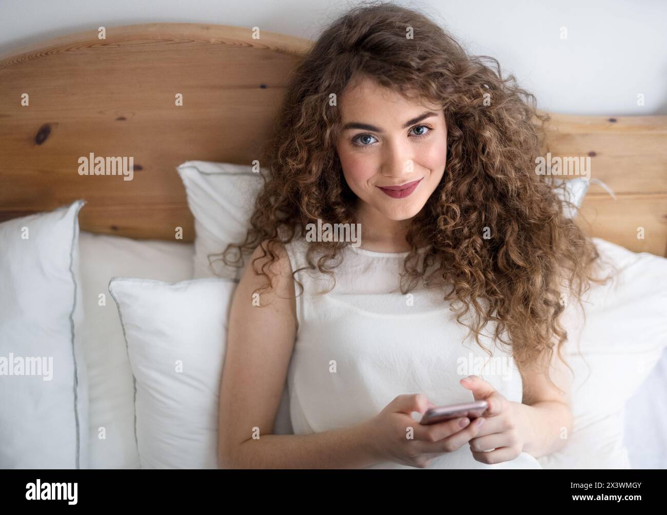 Beautiful woman with curly hair sitting on bed, scolling on smartphone. Stock Photo