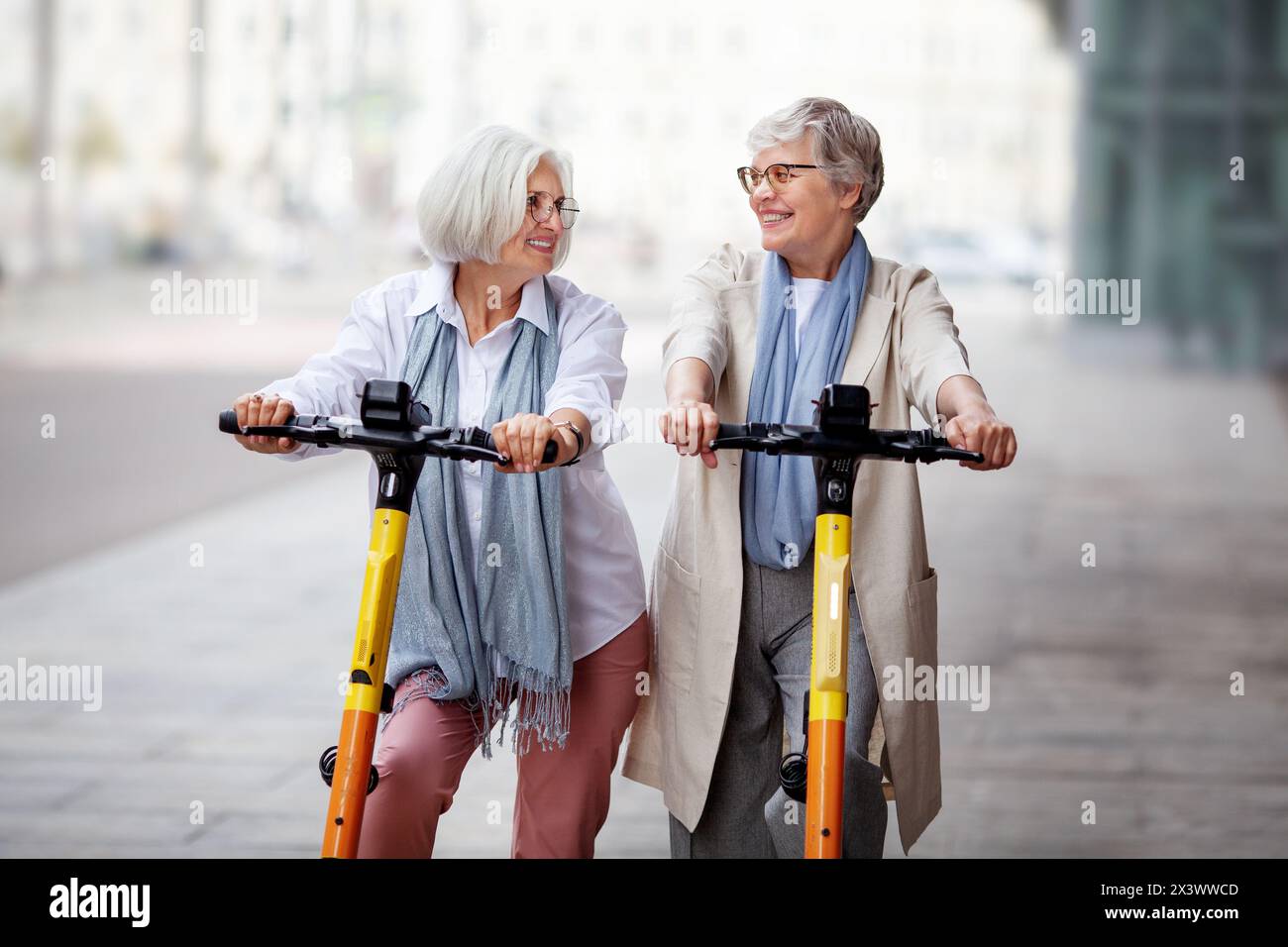 Mature joyful gray haired women friends ride electric scooters in the city, happy smiling, enjoying a fun time together. Stock Photo