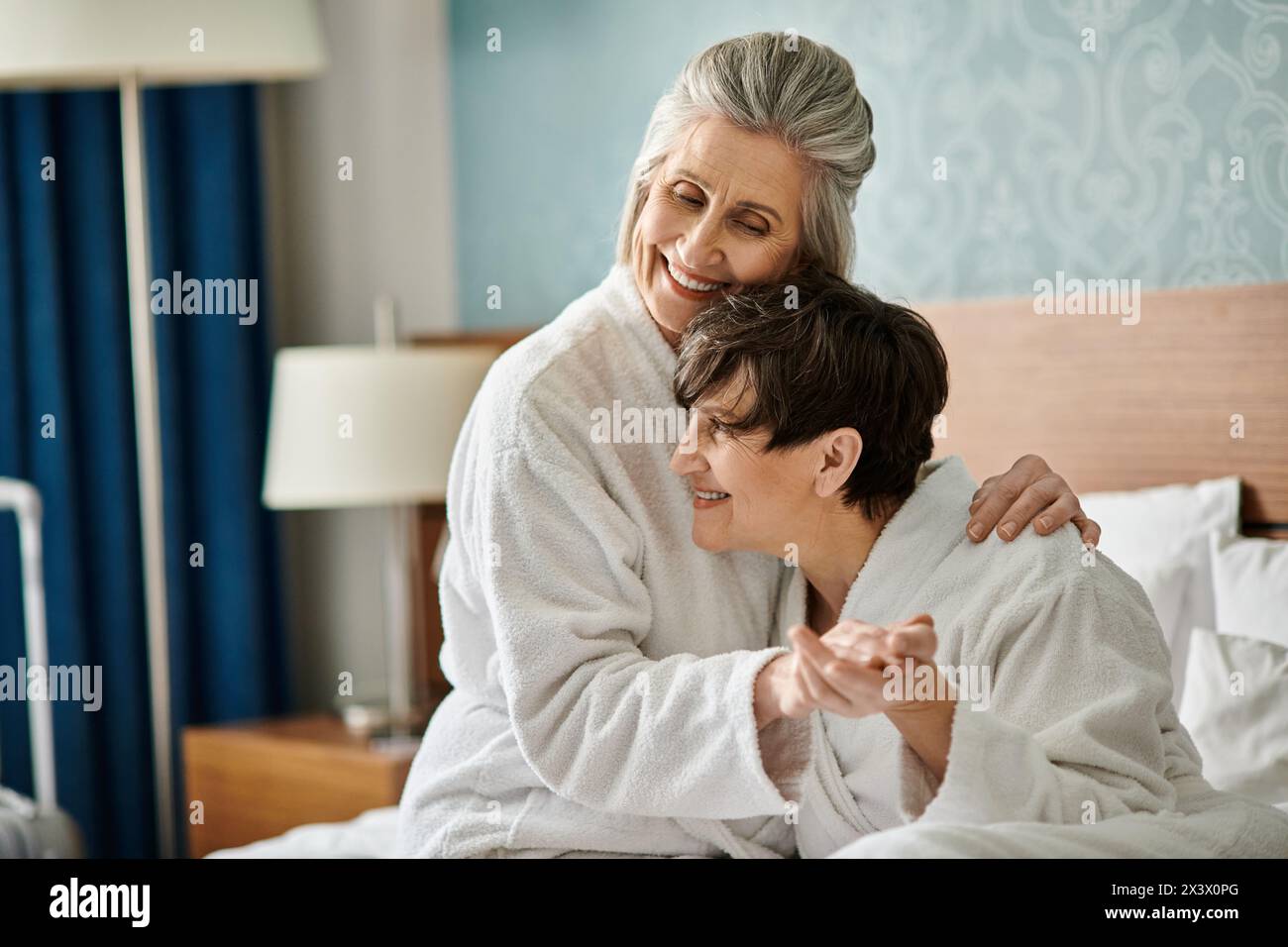 A senior lesbian couple sharing a tender moment in a bed, with a woman in a white robe embracing her partner. Stock Photo