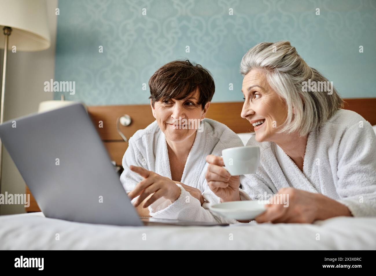 Two women, senior lesbian couple, sit on bed, engrossed with laptop screen in tender moment. Stock Photo