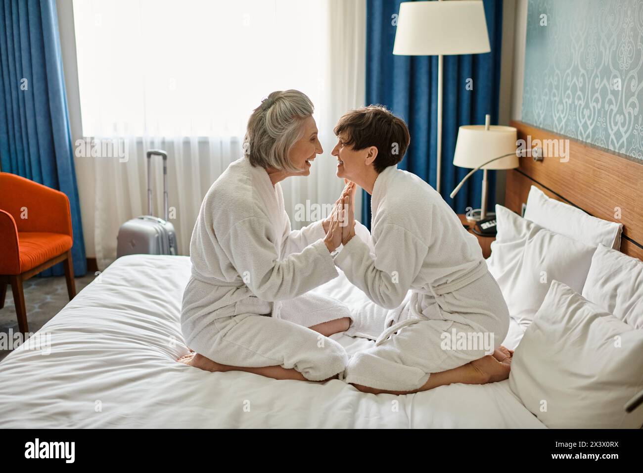 Senior lesbian couple peacefully sitting together on top of a bed in a hotel room. Stock Photo