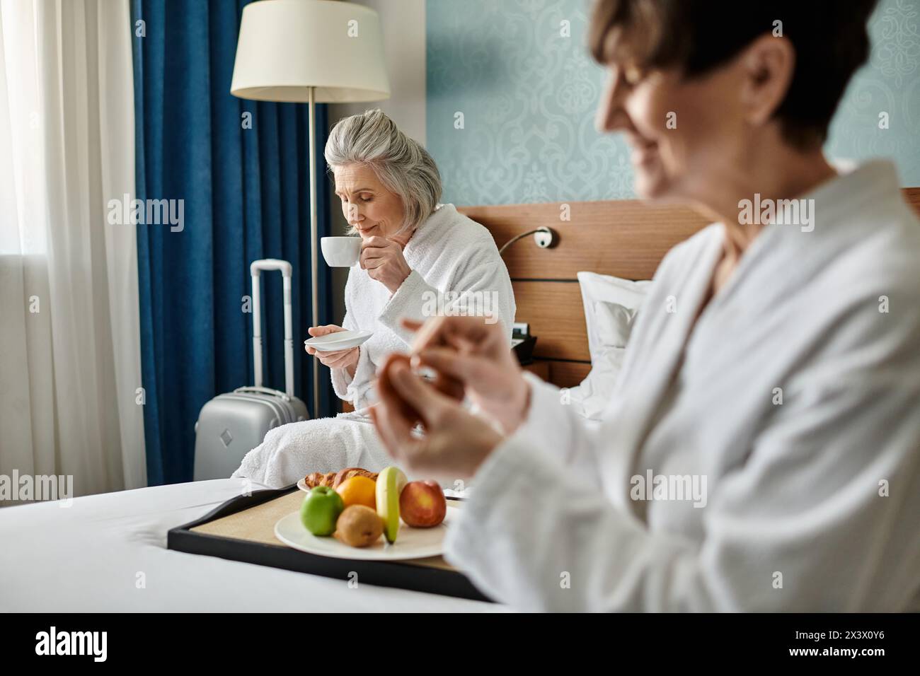 Two senior lesbian women in white robes, tenderly seated on a bed. Stock Photo