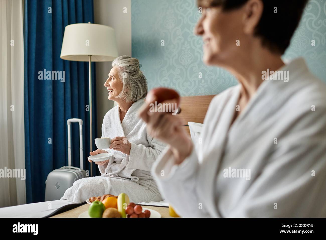 Two senior women in a loving embrace, sitting together on top of a bed in a hotel room. Stock Photo