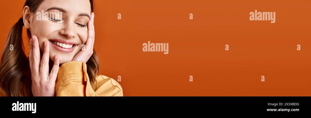 A young, stylish woman in her 20s with hands on face, deep in thought, against an orange studio background. Stock Photo