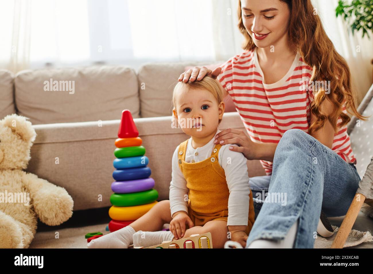 A young mother sits on the floor combing her toddler daughters hair with care and tenderness. Stock Photo