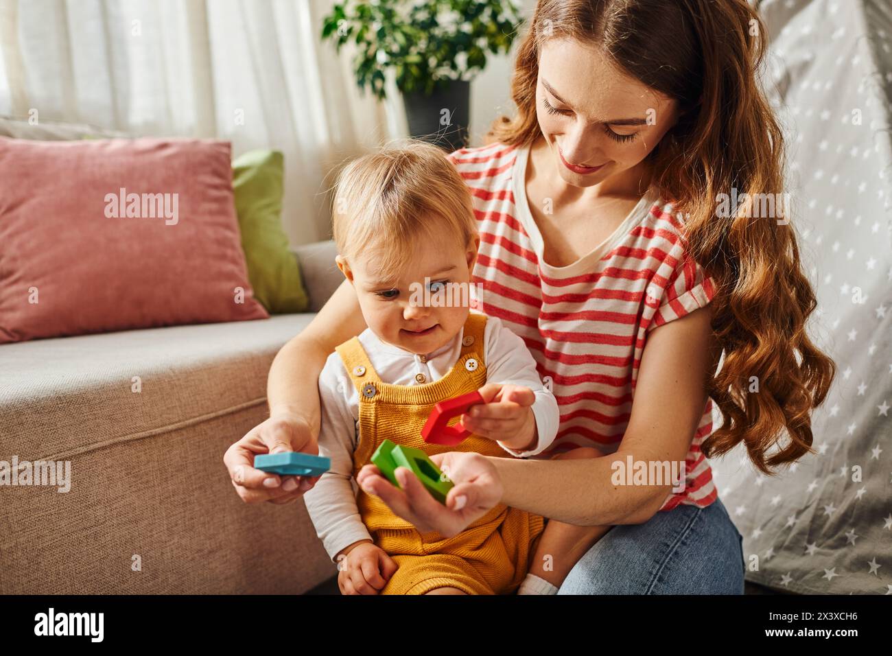A young mother happily interacts with her toddler daughter, engaging in playful activities on the floor at home. Stock Photo