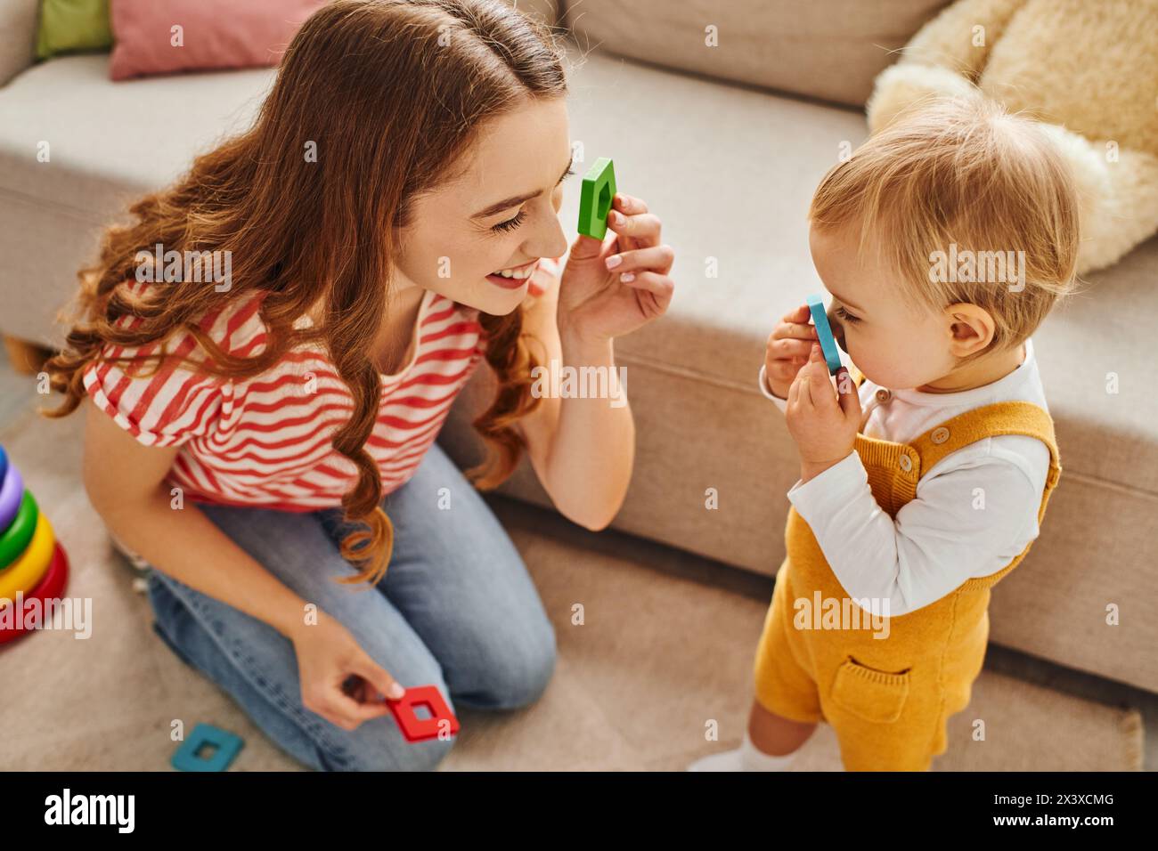 A young mother happily engages with her toddler daughter on the floor, creating a heartwarming moment of joyful interaction. Stock Photo
