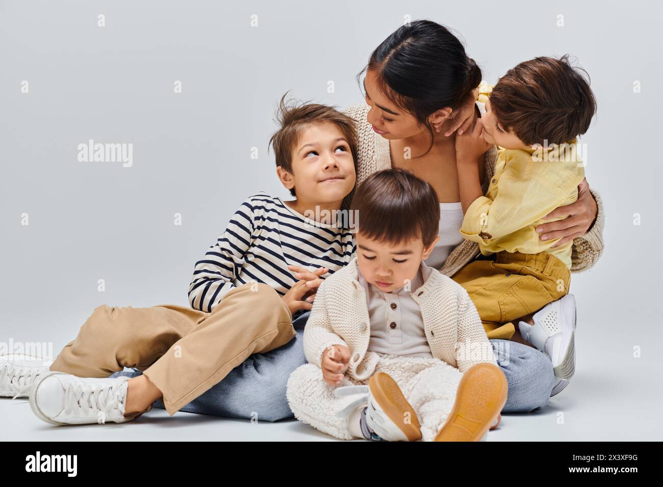 An Asian mother sits on the floor with her children, sharing a loving embrace in a studio against a grey background. Stock Photo