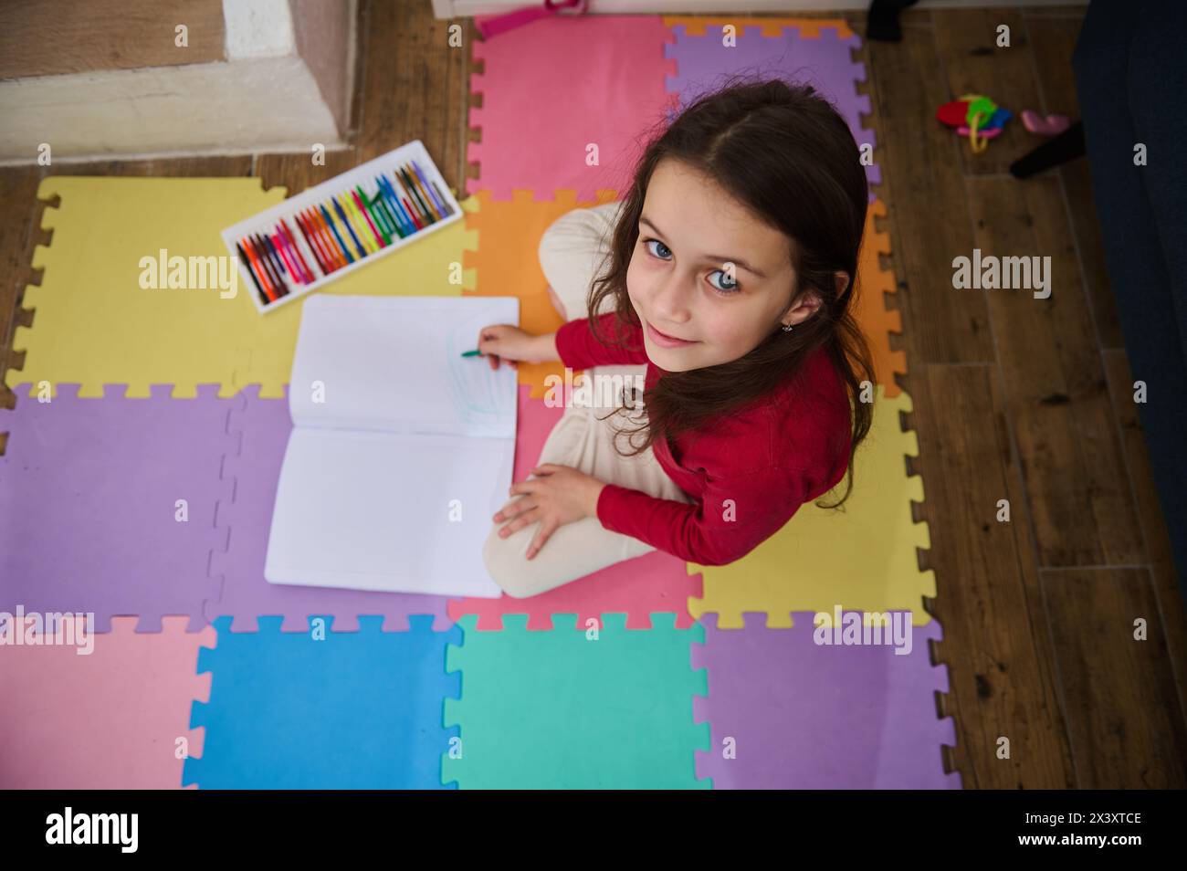 View from above of a cute little child girl smiling looking at camera, drawing with pastel pencils, sitting on a colorful puzzle carpet in cozy domest Stock Photo