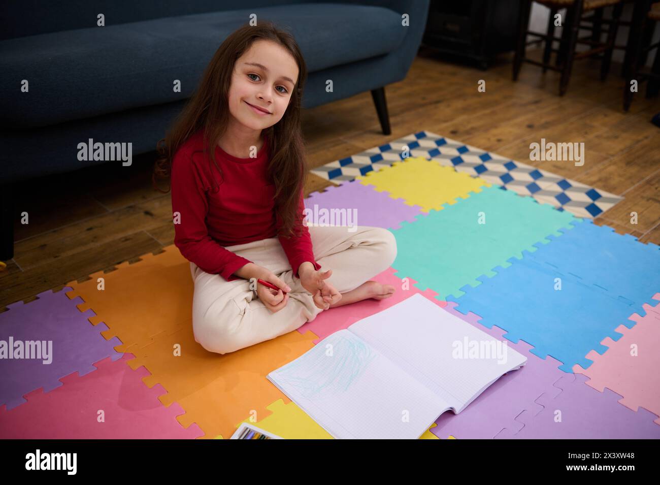 Caucasian lovely little child girl sitting on a colorful puzzle carpet, smiling looking at camera, doing homework. People. Kids. Education concept Stock Photo