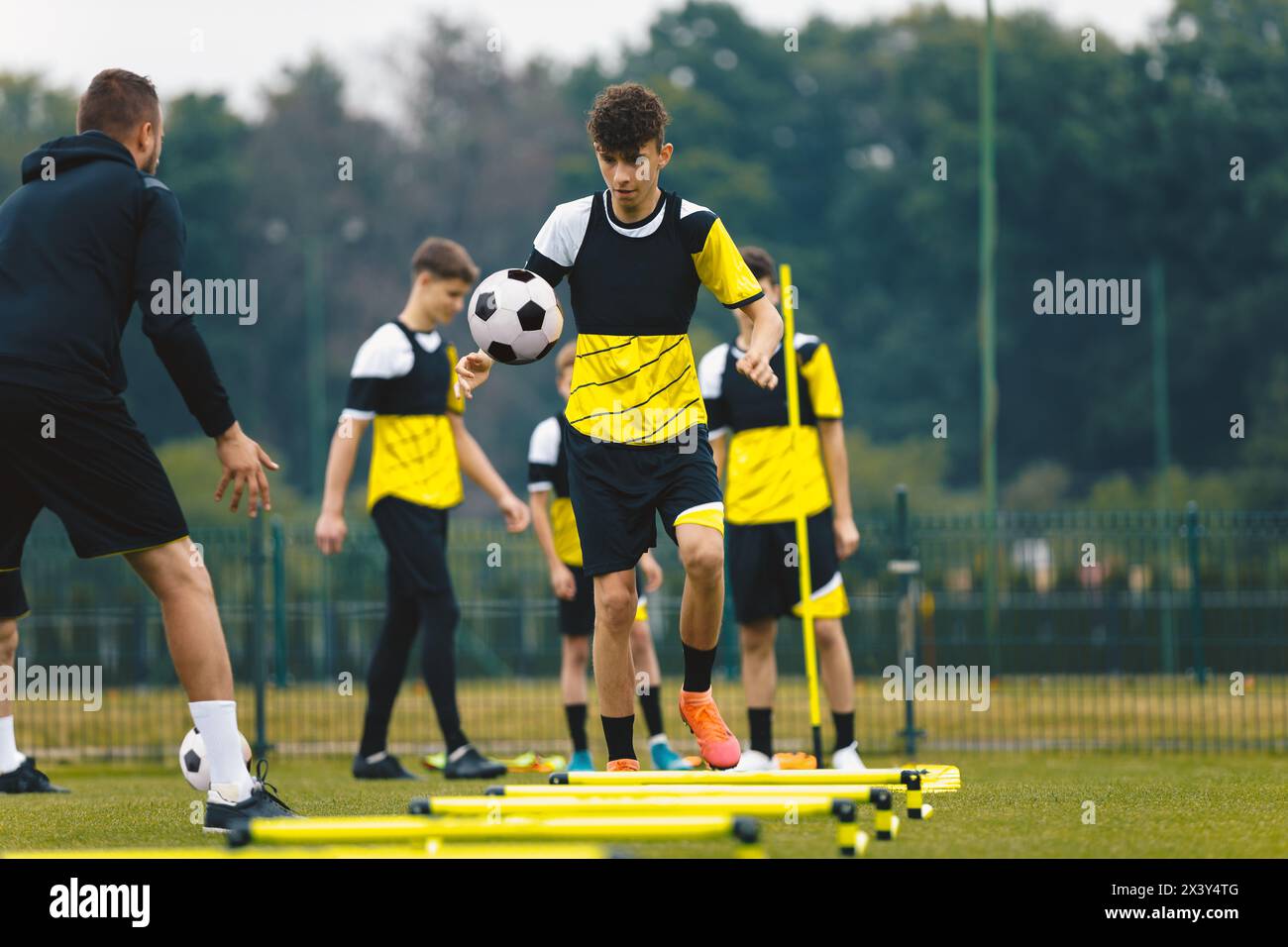 Player kick soccer ball to coach and ladder skipping. Teenagers on soccer training camp. Boys practice football with young coaches. Junior level athle Stock Photo