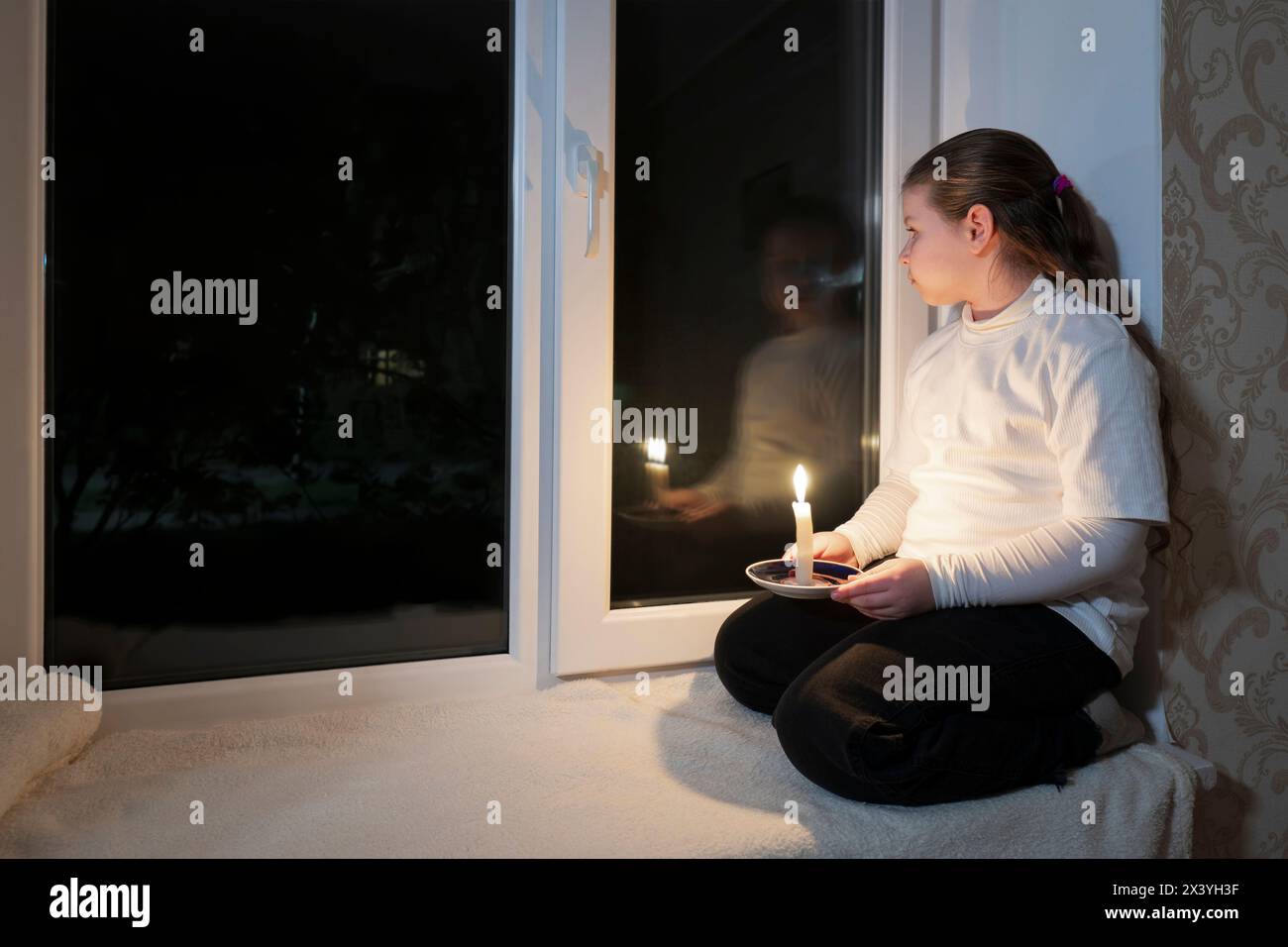 Blackout. Power outage. Girl in a dark room sits on windowsill near window and holds a burning candle in her hands Stock Photo