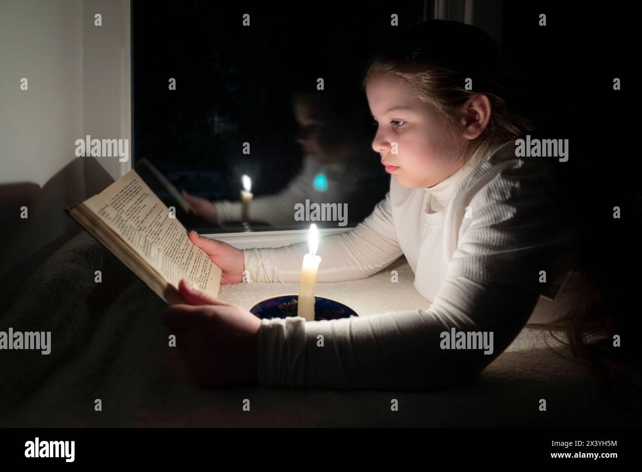 Blackout. A girl in a dark room lies on the windowsill near the window and reads a book by the light of a burning candle. Power outage Stock Photo