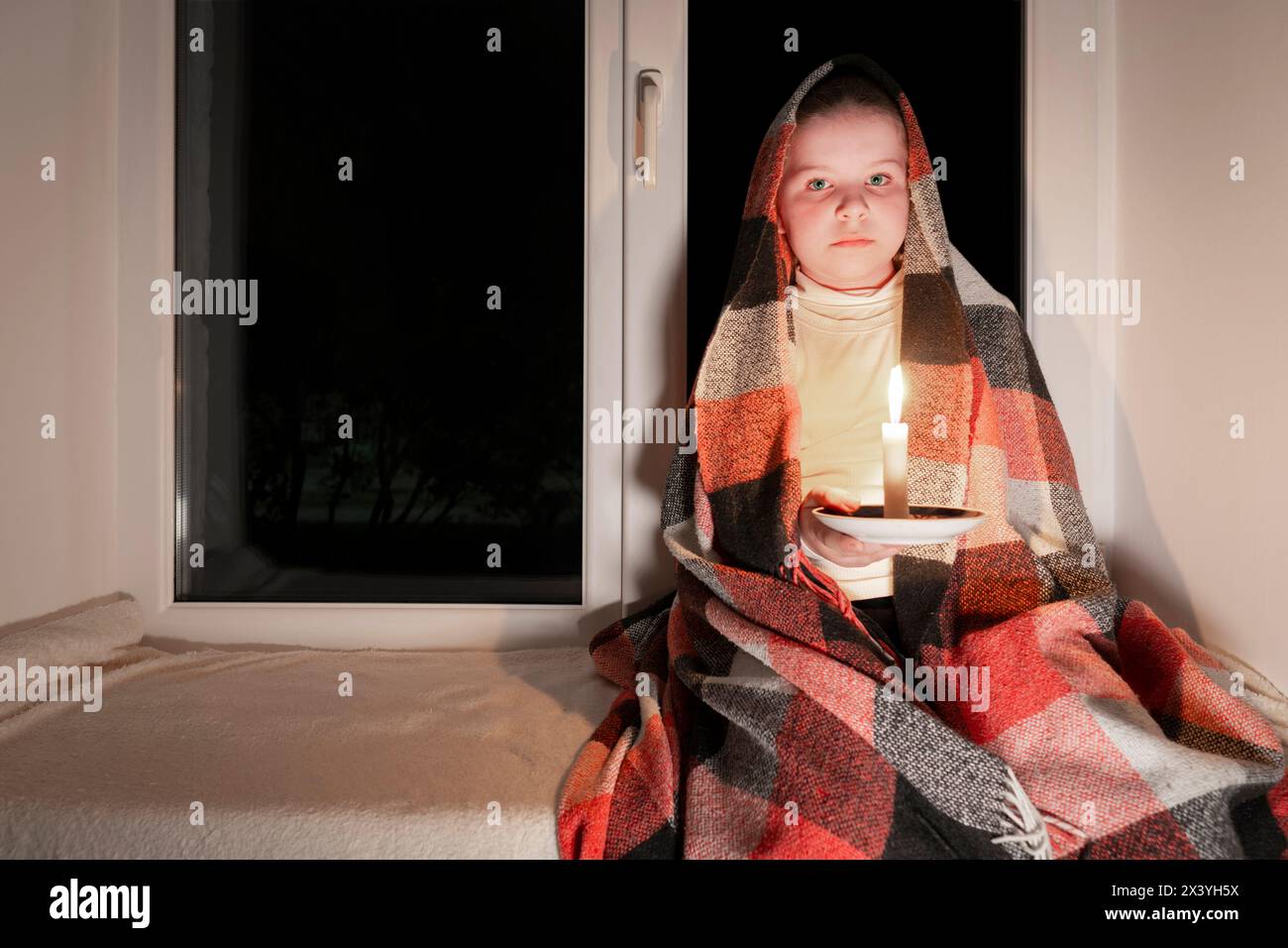 Blackout. A girl, wrapped in a blanket, sits on a windowsill near the window in a dark room and holds a burning candle in her hands. Power outage Stock Photo